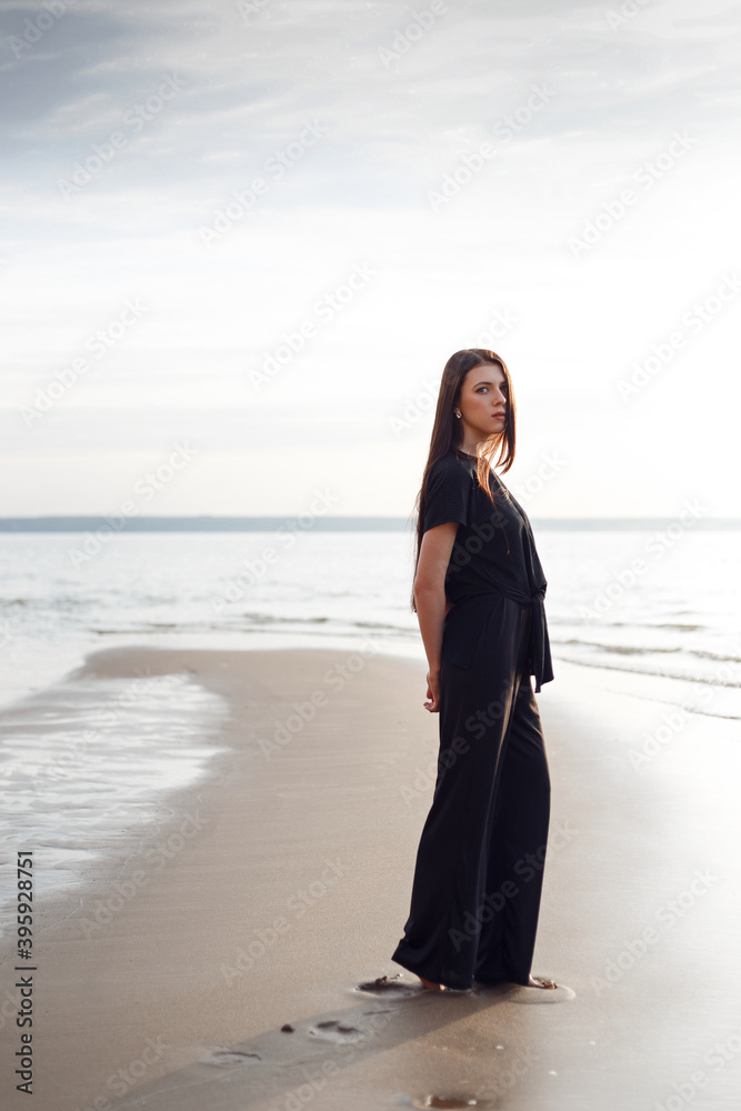 Young beautiful girl in black clothes walking on the beach. She walks along shore. A walk at sunset. Nobody around. Laugh, smile and enjoy summer Vacations. Footprints in the wet sand Young beautiful girl in black clothes walking on the beach. She walks along shore. A walk at sunset. Nobody around. Laugh, smile and enjoy summer Vacations. Footprints in the wet sand