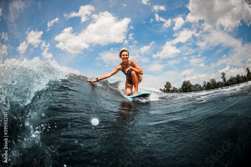 Smiling woman riding on wave sitting on surfboard and touching the water with her hand.