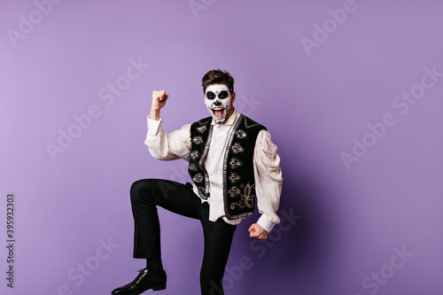 Emotional guy in Mexican clothes rejoices in victory. Photo of man with skull mask posing on lilac background