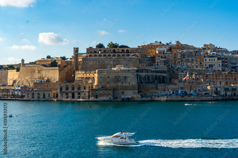 A small yacht in the Grand Harbour in Malta passes the fortified capital city Valletta showing the Lascaris Battery, St. Peter and St. Paul Counterguard, Saluting Battery and Upper Barrakka Gardens.