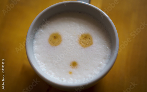 A cup of cappuccino coffee with milk foam showing a thoughtful face, on a blurred wooden table.