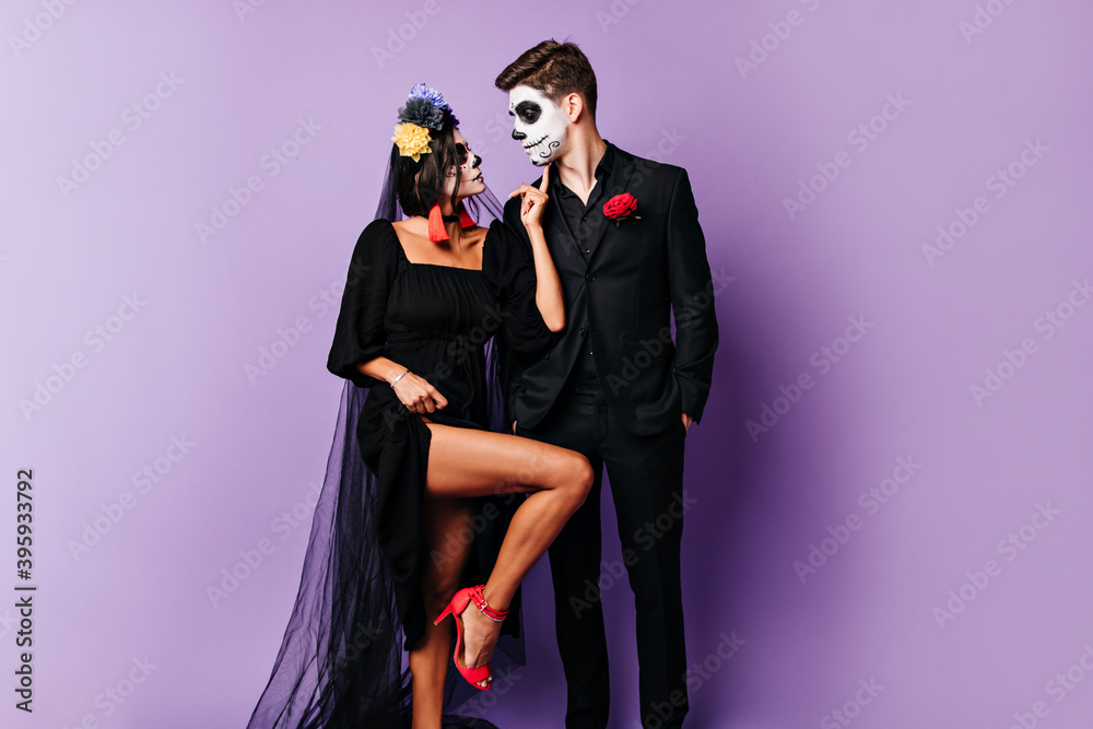 Tanned dark-haired woman in black wedding veil and make-up on maksarad coquettishly raised her leg while her man looking in love at her Tanned dark-haired woman in black wedding veil and make-up on maksarad coquettishly raised her leg while her man looking in love at her