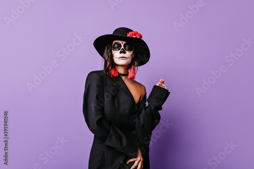 Curly stylish girl with red earrings and rose on black wide-brimmed hat posing pathetically in outfit for Halloween photo