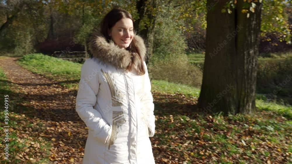 Beautiful woman with brown hair walking in park on cold autumn day with trees covered in yellow leaves in warm fur collar coat Beautiful woman with brown hair walking in park on cold autumn day with trees covered in yellow leaves in warm fur collar coat
