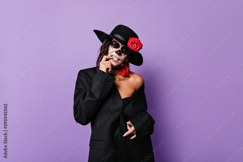 Pathetic woman mafioso in black outfit touches her teeth and poses in skull mask on lilac isolated background Pathetic woman mafioso in black outfit touches her teeth and poses in skull mask on lilac isolated background