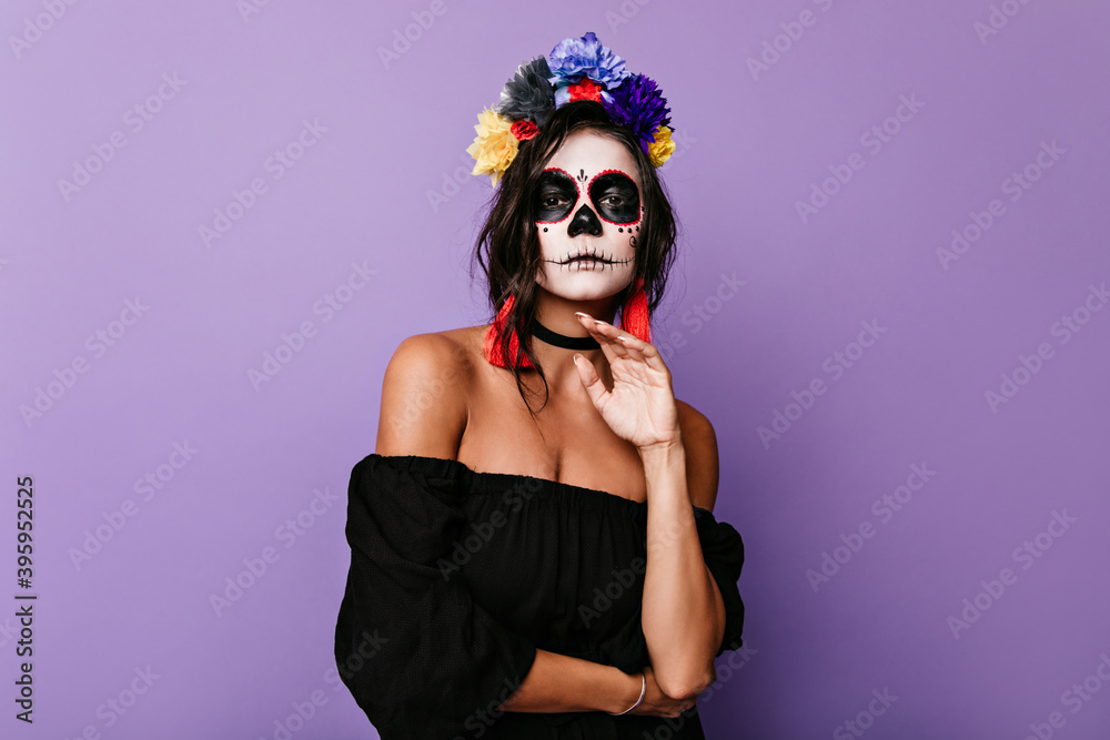Attractive brunette with mexican mask poses in halloween outfit. Woman in black dress confidently looks into camera Attractive brunette with mexican mask poses in halloween outfit. Woman in black dress confidently looks into camera