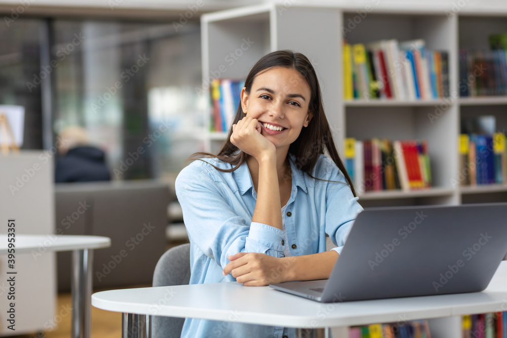 Beautiful female student or female employee sitting at table in the university library or office with laptop looking at the camera. Remote work or education concept Beautiful female student or female employee sitting at table in the university library or office with laptop looking at the camera. Remote work or education concept
