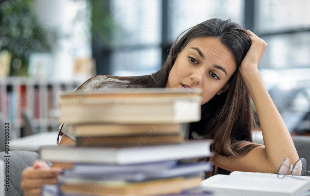 Beautiful frustrated female student looks at stack of books and holds her head while sitting at table in the university library. Studying and education concept Beautiful frustrated female student looks at stack of books and holds her head while sitting at table in the university library. Studying and education concept