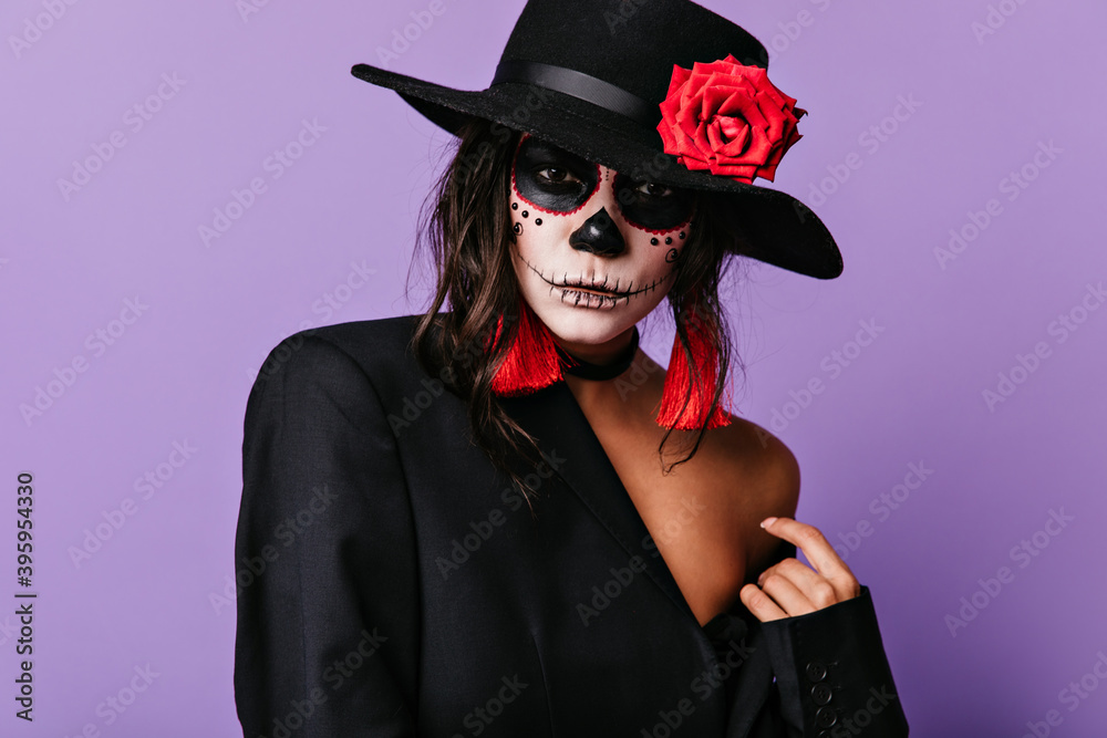 Attractive brunette in hat with red rose and painted face is looking confidently at camera. Closeup portrait of woman with bright accessories Attractive brunette in hat with red rose and painted face is looking confidently at camera. Closeup portrait of woman with bright accessories