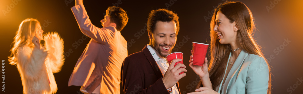 Φωτογραφία woman and african american man holding plastic cups near friends during party o