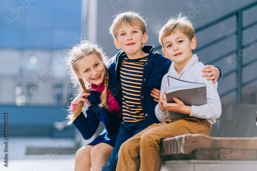 Photography Three cute blond little children, two boys and girl with book hugging while sitting on the bench in the schoolyard at the sunny day
