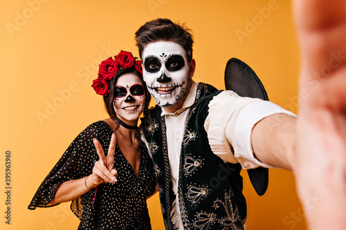 Joyful guy and girl are enjoying halloween party. Couple takes selfie in unusual clothes showing peace sign