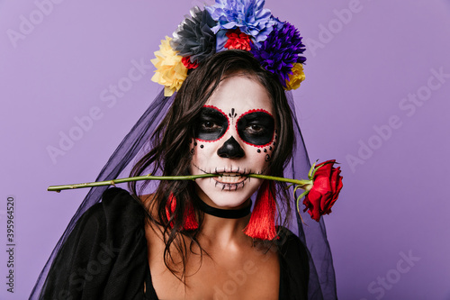 Passionate Mexican woman with painted face holding red rose in her teeth. Closeup photo of curly brunette with colorful flowers in her hair