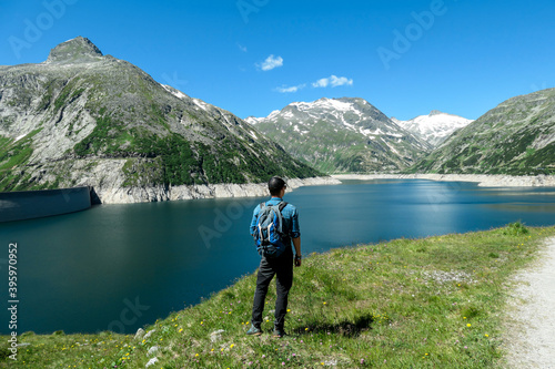 Man standing at the side of a lake at Kölnbreinsperre dam in Austria. The lake has navy blue color. High Alps around. There is a glacier in the back. The meadows blossoming with wild flowers. Calmness
