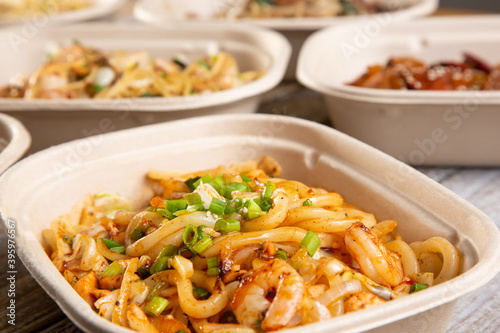 A closeup view of a fast casual bowl featuring fat Chinese noodles and shrimp, among several other entrees on a table.