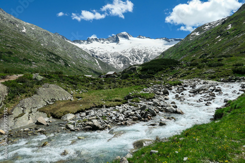 Panoramic view on huge glacier in Maltal region in Austrian Alps. The valley below it flourishing with green and freshness., small torrent in the middle. Summer in the mountains. Serenity and solitude