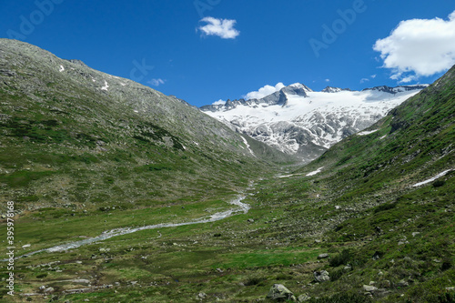 Panoramic view on huge glacier in Maltal region in Austrian Alps. The valley below it flourishing with green and freshness., small torrent in the middle. Summer in the mountains. Serenity and solitude
