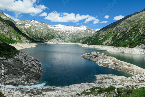 A torrent rushing towards a lake. The artificial lake stretches over a vast territory, shining with navy blue color. The dam is surrounded by mountains.A glacier in the back. Controlling the nature