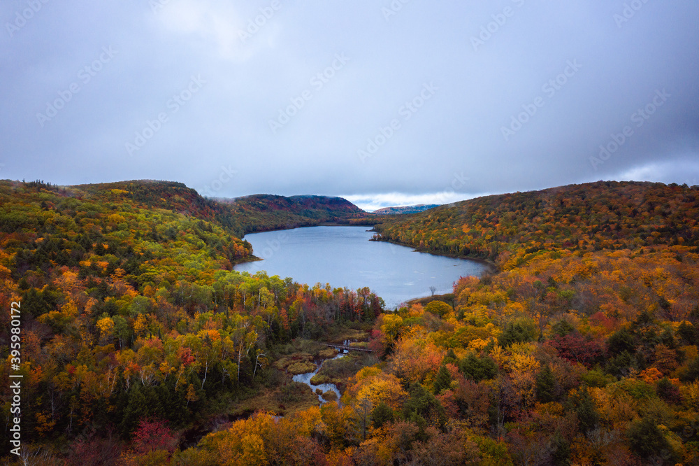 Beautiful travel aerial photograph of Lake of the Clouds and the pedestrian bridge over Carp River on an overcast morning with colorful autumn foliage blanketing the hills and valley below.