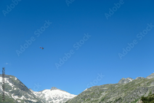 A rescue helicopter flying over the Alps in Austria. Rescue mission in a difficult terrain. There are high, snow capped mountains around. The helicopter flies high above the peaks