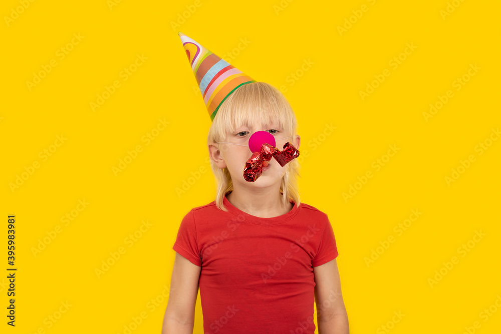 Child in festive hat with red clown nose and whistle. Festive mood. Portrait of boy on yellow background