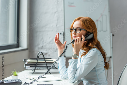  businesswoman gesturing and talking on landline telephone, while sitting at workplace on blurred background photo