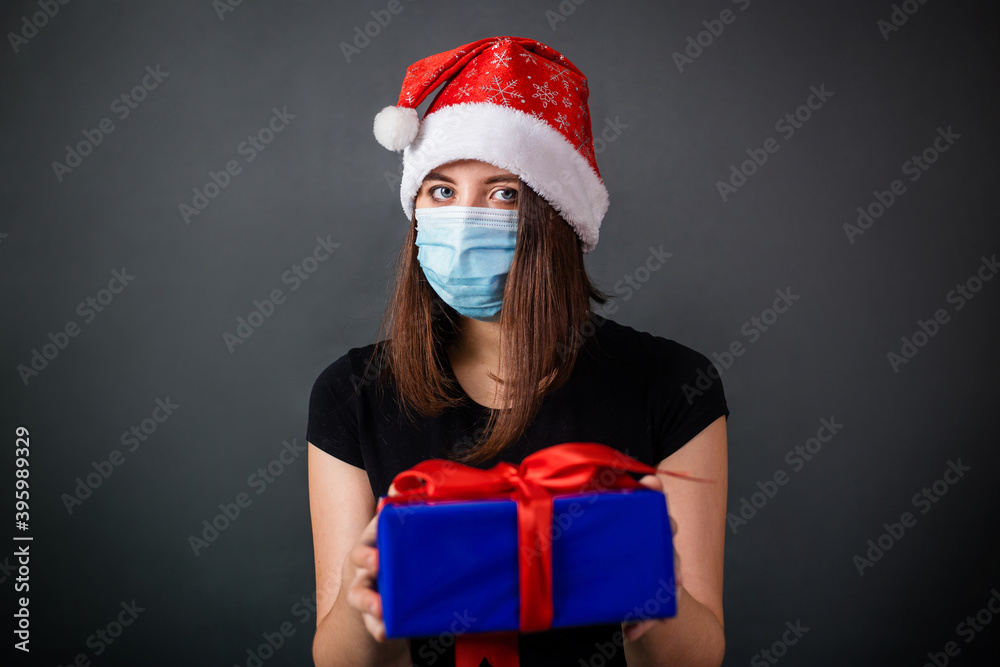 A woman in a Christmas cap and a medical mask holds a gift box in her hands while standing against a gray background. A woman in a Christmas cap and a medical mask holds a gift box in her hands while standing against a gray background.