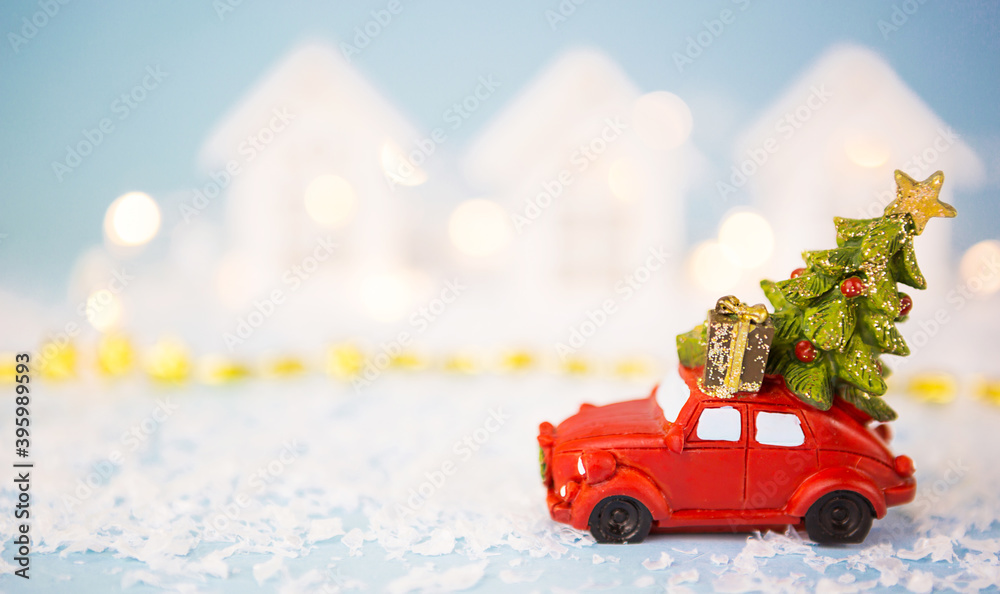 Christmas decor - red retro car on snow carries past white houses with lights garlands in bokeh Christmas tree with gift boxes on roof. Toy on blue background, space for text. New Year in town