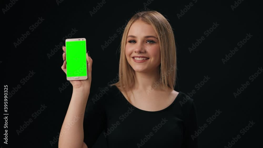 Happy blonde standing over black background and holding modern smartphone with green empty screen. Concept of people and technology.