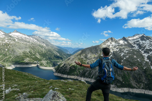 Man with hiking backpack admiring a view on an artificial lake at a dam in Austria from above. The lake has navy blue color. High Alps around. There is a glacier in the back. Adventure and discovery