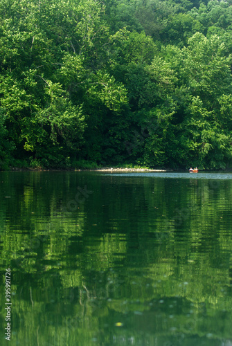 Majestic vertical nature background woth two tiny kayakers in the distance. Green foliage reflected in river water for beautiful tranquil effect in bright sunshine with copy space.