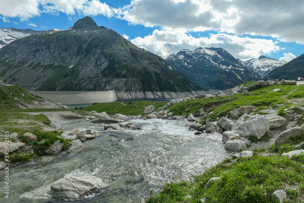 A small torrent in the Austrian Alps flowing to the lake. The meadow around it is overgrown with lush green grass. In the back there is a glacier. Sunny and bright day. Power of the nature. Remedy