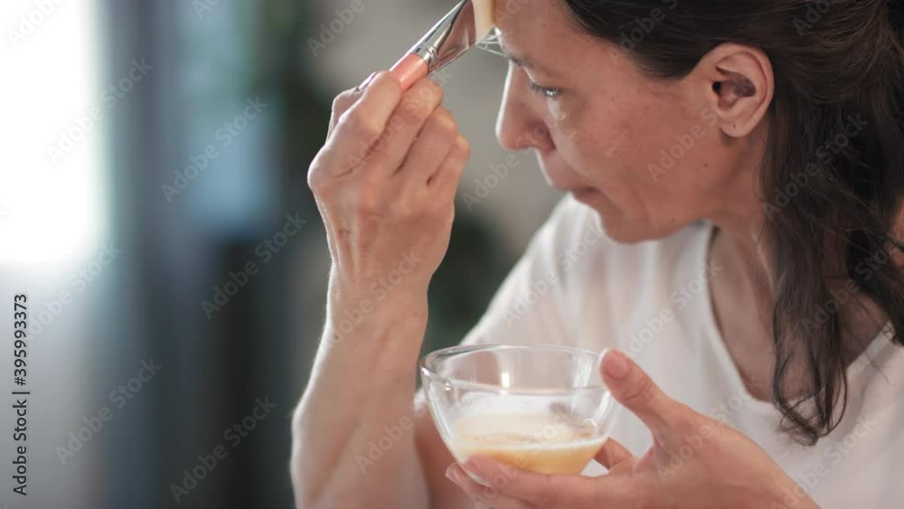 Close up of groomed woman applying liquid egg mask to forehead with fan brush looking at mirror taking care of her skin at home Close up of groomed woman applying liquid egg mask to forehead with fan brush looking at mirror taking care of her skin at home