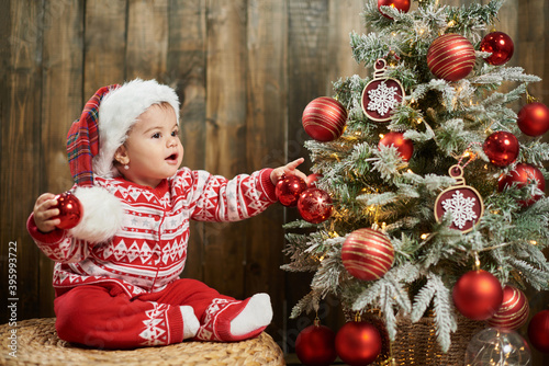 Adorable girl child wearing red sweater next to a christmas tree against wooden wall