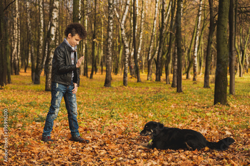 A curly-haired brown-haired boy with a mohawk hairstyle in a leather jacket on the background of an autumn forest plays with a dog.