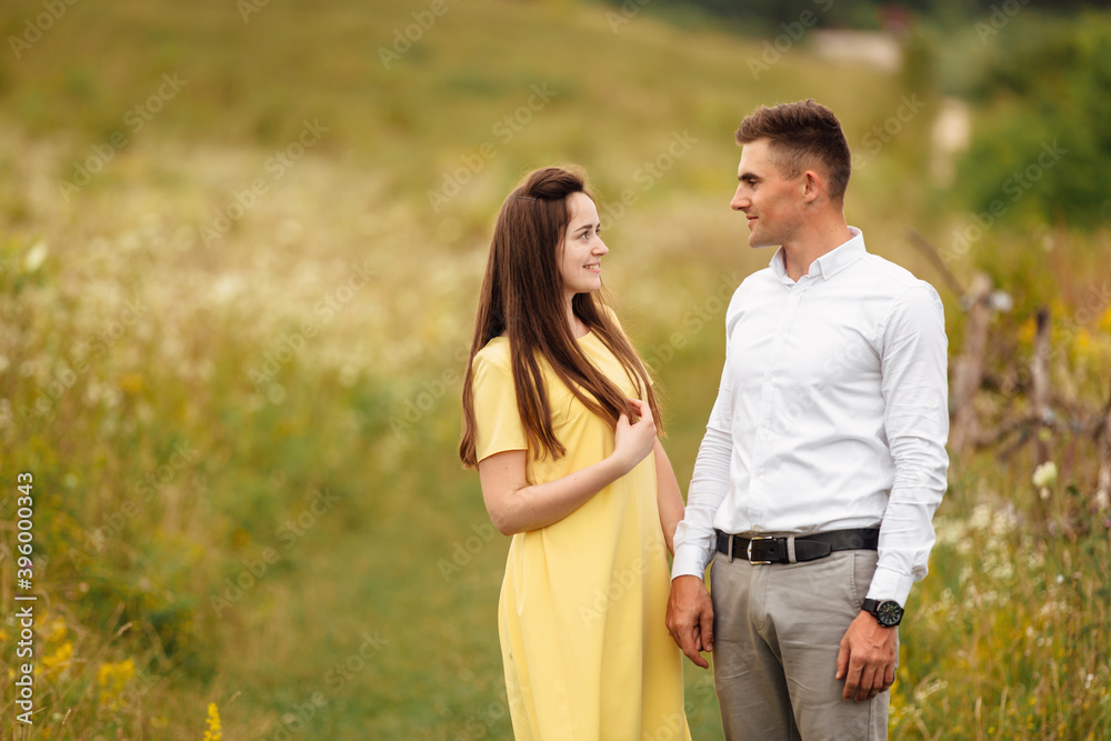 young couple in love look at each other in a grass field in sunny day. girl in a yellow dress and a guy with a stylish haircut. portrait of couple walking on countryside young couple in love look at each other in a grass field in sunny day. girl in a yellow dress and a guy with a stylish haircut. portrait of couple walking on countryside