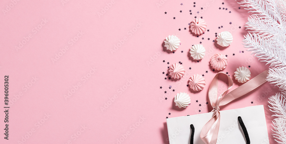 Festive Christmas and New Year banner with white paper bag with a bow, sweet meringue cookies, shining decorations and fir tree branches on light pink background with copy space for your text. Festive Christmas and New Year banner with white paper bag with a bow, sweet meringue cookies, shining decorations and fir tree branches on light pink background with copy space for your text.