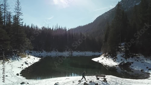 Drone shot of a man walking around the shore of Green Lake, Austrian Alps. The lake shimmers with many shades of green and turquoise. Winter in the mountains. There is snow on the ground. Wandering
