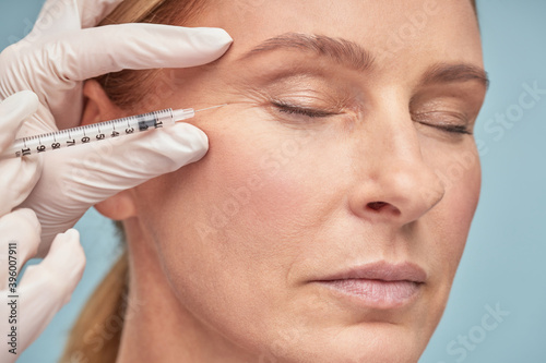 Close up portrait of a beautiful mature woman keeping eyes closed while receiving hyaluronic acid injection by beautician photo