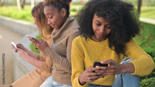 A young black woman is leafing through a social network or looking at photos on her phone, her friends are sitting next to her and also using phones at this time