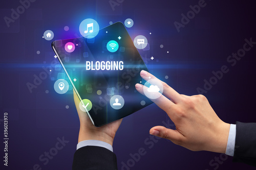 Businessman holding a foldable smartphone with BLOGGING inscription, social media concept