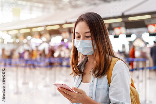 Young woman wearing a face mask and holding a mobile phone is looking for a check-in counter at the airport, New normal lifestyle concept
