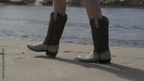 A girl in a white dress and cowboy boots walks on a sandy beach. Focus on your feet.