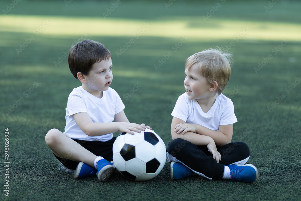 Lifestyle of two children and soccer ball, sitting on grass, laughing and talking. Concept about childhood, sports and communication of preschool.