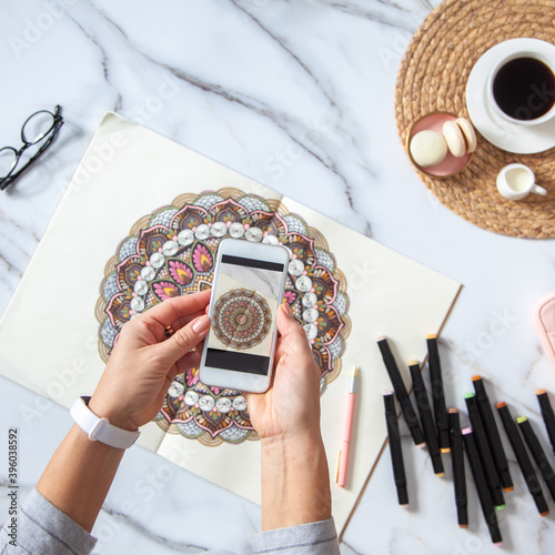 Photography Woman is taking photos of colored mandala from sketchbook on white marble desk w