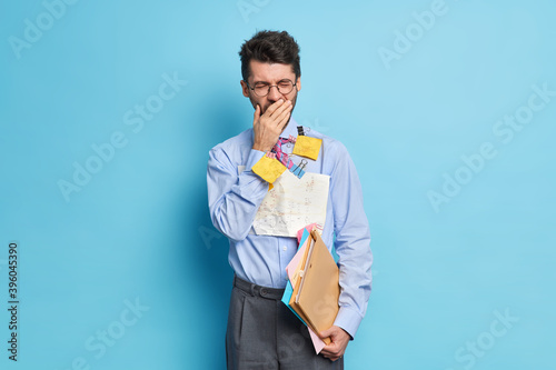 Fotografie Photo of exhausted man yawns after long hours of working prepares financial report wears formal clothes poses indoor against blue background