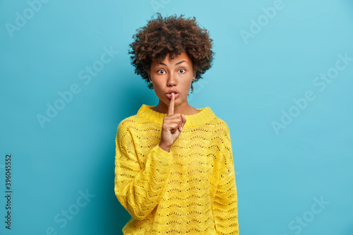 Obraz na płótnie Surprised dark skinned young woman makes shush gesture asks to keep information in secret shows hush sign has shocked face expression wears casual yellow sweater isolated over blue background