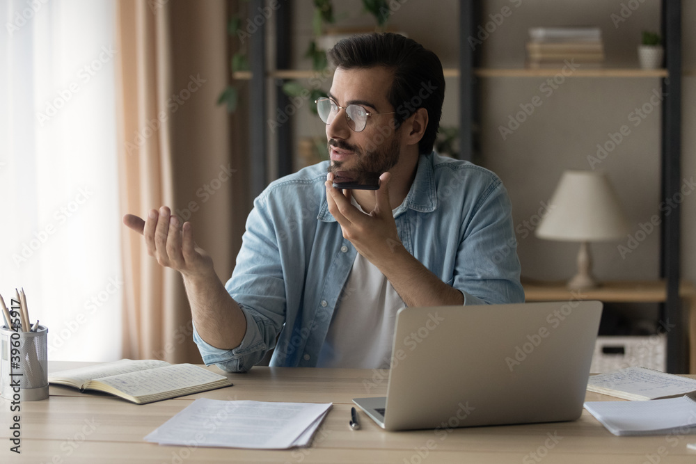 Young Caucasian man sit at desk in home office work on laptop record audio message on smartphone. Millennial male use computer speak talk on call activate digital voice assistant on cellphone. Young Caucasian man sit at desk in home office work on laptop record audio message on smartphone. Millennial male use computer speak talk on call activate digital voice assistant on cellphone.