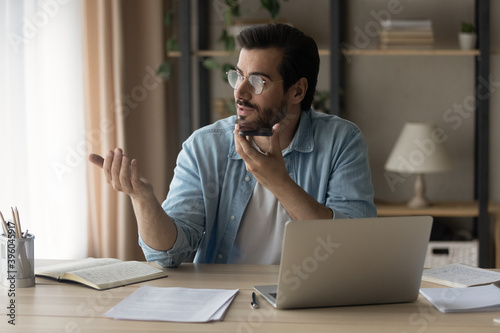 Young Caucasian man sit at desk in home office work on laptop record audio message on smartphone. Millennial male use computer speak talk on call activate digital voice assistant on cellphone.