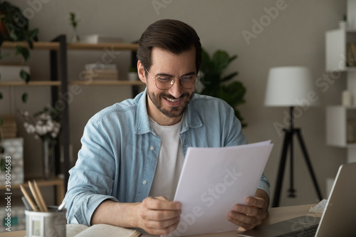 Happy millennial Caucasian man sit at desk at home office read good news in post paper letter. Smiling young male feel excited get pleasant message or notice in postal paperwork or correspondence.